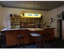Kleine, gemütliche Bar mit U-förmigem Tresen aus Holz und weißer Oberfläche. Im Hintergrund befindet sich ein beleuchtetes Regal mit verschiedenen Getränkeflaschen, Gläsern und Dekor, darüber ein Schild mit der Aufschrift „Kindl-Pils“.