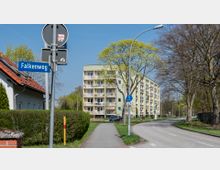 Mehrstöckiges Wohngebäude mit Balkonen im Hintergrund, umgeben von Bäumen und Grünflächen. Im Vordergrund führen eine Straße und ein Geh- und Radweg vorbei, neben einem Straßenschild mit der Aufschrift „Falkenweg“.