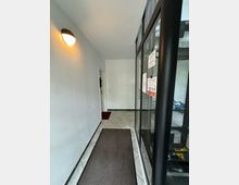 Eingangsbereich eines Gebäudes mit weißen Wänden, Fliesenboden und einem braunen Teppichläufer. Rechts befindet sich eine Glaswand mit schwarzen Rahmen, links eine Wandlampe, und am Ende des Flurs eine geschlossene Tür.