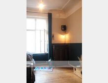 Ein schlicht eingerichtetes Zimmer mit Holzfußboden und hellbraunen Wänden. Ein großes Fenster mit Vorhängen lässt Tageslicht herein, daneben steht ein dunkler Holzschrank mit einer Glasflasche darauf; an der Wand hängt ein kleiner Lautsprecher.