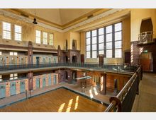 Ehemaliges Schwimmbad in einem historischen Gebäude mit hohen Decken, großen Fensterfronten und Backsteinwänden. Der Raum zeigt zwei Ebenen mit Umkleidekabinen und eine leere Schwimmhalle mit einem kleinen Sprungbrett.