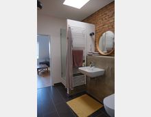 Modernes Badezimmer mit dunklem Fliesenboden, einer Ziegelwand, einem weißen Waschbecken und einem runden Spiegel. Eine Dusche mit Glastür, ein Handtuchwärmer mit aufgehängten Handtüchern und ein gelber Teppich auf dem Boden sind ebenfalls sichtbar.