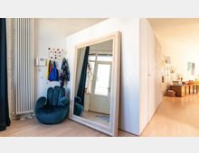 Innenraum mit modernem Design; ein großer Standspiegel mit Holzrahmen lehnt an einer weißen Wand neben einem türkisfarbenen Sessel in Form einer Hand. Im Hintergrund ist eine Glastür zu sehen, und rechts befindet sich ein Holzschrank mit Dekorationen auf einer hellen Holzfußbodenfläche.