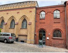 Backsteinfassade eines Gebäudes mit gotischen Fenstern, bestehend aus gelbem und rotem Ziegelstein. Links steht ein geparktes Auto vor der gelben Backsteinseite, rechts zeigt die rote Fassade einen Eingang mit Rundbogenfenstern.