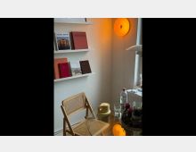 Ein kleiner Raum mit minimalistischer Einrichtung: An der weißen Wand sind zwei schmale Regalbretter mit Büchern unterschiedlicher Farben angebracht, daneben eine runde orangefarbene Wandlampe. Im Vordergrund stehen ein geflochtener Holzstuhl und ein Glastisch mit Wasserflasche, Gläsern und Dekoration.