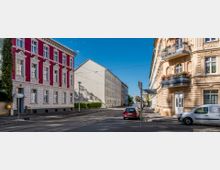 Straßenszene mit mehrstöckigen Wohnhäusern im Altbaustil; links ein rotes Gebäude mit weißen Fensterelementen, rechts ein beiges Gebäude mit Balkons und Blumenkästen. Die Straße ist mit parkenden Autos gesäumt, und Straßenbahnschienen verlaufen mittig.