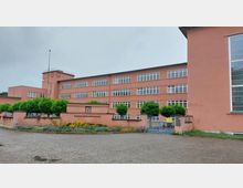 Rosa Schulgebäude der Friedrich-Ebert-Grundschule mit mehreren rechteckigen Fenstern, einer zentralen Turmstruktur mit Uhr und umliegenden gepflegten grünen Bäumen und Blumenbeeten. Im Vordergrund ein gepflasterter Platz und ein blaues Metalltor.