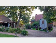 Ein idyllischer Hof mit Kopfsteinpflaster, einem kleinen Garten mit Blumen und Sträuchern, einem Holzunterstand und einem grauen Bauwagen. Im Hintergrund sind ein Ziegelgebäude mit rotem Dach und eine Fachwerkstruktur sichtbar, umgeben von großen Bäumen und ländlicher Vegetation.