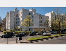 Wohngebäude aus Beton mit moderner, kantiger Architektur, flankiert von Bäumen und einer Reihe geparkter Autos entlang der Straße. Im Vordergrund sind Gehwege, ein Fußgänger und ein kleiner Grünstreifen zu sehen.