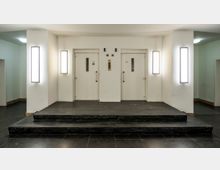 Weißer Aufzugsvorraum mit zwei Aufzugstüren nebeneinander, flankiert von wandmontierten vertikalen Leuchten. Der Boden ist aus dunklem Stein, und die Aufzüge sind über eine kleine Stufe erreichbar.