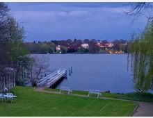 Blick auf einen See in einer Parklandschaft bei Abenddämmerung, mit einem kleinen Steg und weißen Bänken im Vordergrund. Im Hintergrund sind beleuchtete Gebäude am gegenüberliegenden Ufer zu sehen, umgeben von Bäumen.