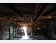 Ein leerer Dachboden mit sichtbaren Holzbalken und einem offenen Ziegeldach, durch das Sonnenlicht einfällt. An der linken Seite befindet sich eine grüne Holztür, und der Boden ist teilweise mit Schutt bedeckt.