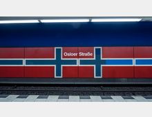 U-Bahn-Station mit dem Namen "Osloer Straße", Wandgestaltung in den Farben Rot, Blau und Weiß mit Kreuzmustern, die an die norwegische Flagge erinnern. Im Vordergrund sind Gleise und ein gepflasterter Bahnsteig zu sehen.
