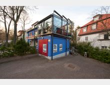 Zweigeschossiges Gebäude mit blauer Fassade, einer roten Tür und einem verglasten Obergeschoss. Das Gebäude befindet sich in einer baumbestandenen Umgebung mit gepflastertem Vorplatz und ist an ein rotdachiges Wohnhaus angrenzend.