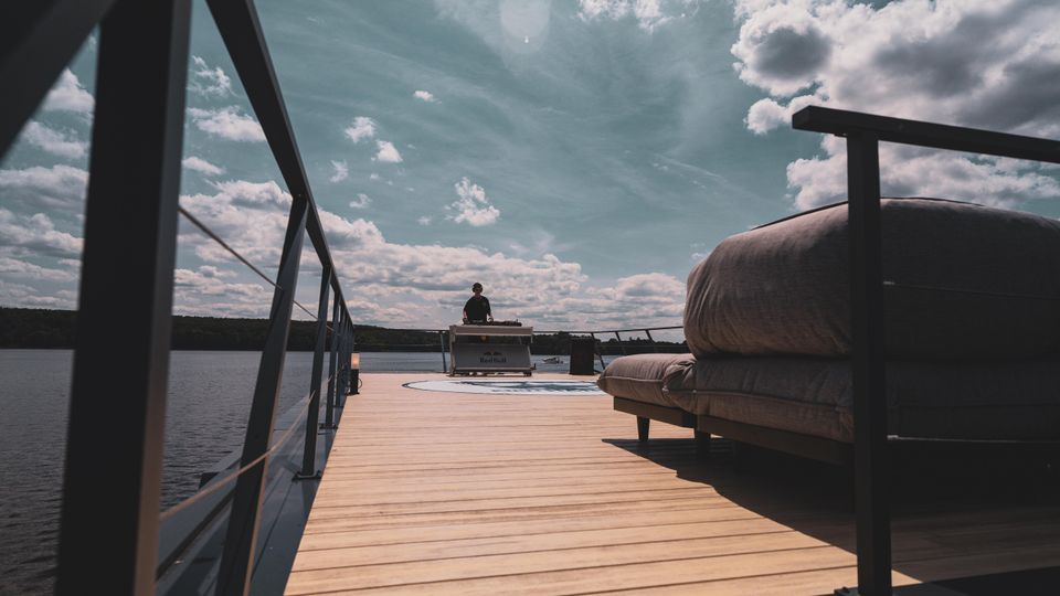Eine offene Holzplattform auf einem See, umgeben von einem Geländer, mit einem Sofasitzbereich auf der rechten Seite und einer DJ-Station in der Mitte. Der Himmel ist leicht bewölkt, und im Hintergrund erstreckt sich eine bewaldete Uferlinie.
