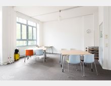 Heller Seminarraum mit weißen Wänden, großen Fenstern, grauem Teppichboden und einfachen Tischen und Stühlen in verschiedenen Farben. Im Raum befinden sich zusätzlich gestapelte Klapptische und eine Uhr an der Wand.