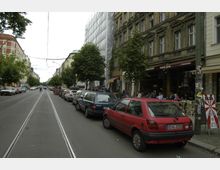 Straßenszene in einer Stadt mit geparkten Autos entlang beider Seiten der Straße, Straßenbahngleisen in der Mitte und Häusern mit mehreren Stockwerken im Hintergrund. Auf der rechten Seite befinden sich ein Café mit Sitzplätzen im Freien und Bäume entlang des Gehwegs.