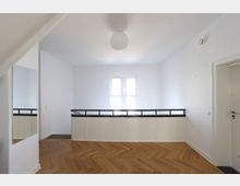 Minimalistischer Innenraum mit weiß gestrichenen Wänden, einem Fenster mit milchigem Glas in der Mitte und einem Parkettboden in Fischgrätenmuster. Links befindet sich ein großer Spiegel, rechts eine weiße Tür und davor ein Geländer mit schwarz-weißer Gestaltung.