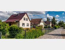 Eine Reihe von Einfamilienhäusern mit Satteldächern und gepflegten Vorgärten, die durch niedrige Metallzäune abgegrenzt sind, entlang eines gepflasterten Gehwegs in einer ruhigen Wohnstraße. Im Hintergrund sind Bäume und ein teilweise bewölkter Himmel zu sehen.