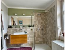 Modernes Badezimmer mit wandhängendem Holzwaschbecken, einem großen Spiegel, einer offenen Dusche mit Glasabtrennung und beigefarbenen Fliesen. Rechts befindet sich ein großes Fenster, das natürliches Licht hereinlässt.