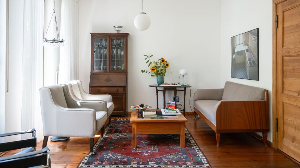 Helles Wohnzimmer mit Holzdielenboden, großem Fenster mit weißen Vorhängen und einem gemusterten Teppich unter einem niedrigen Couchtisch. Im Raum stehen zwei Sessel, ein Sofa, ein Holzvitrinenschrank sowie ein Beistelltisch mit Blumen; an der Decke hängt eine runde Pendelleuchte.