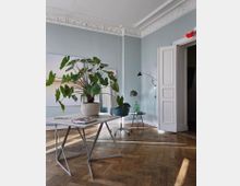 Helles, stilvolles Büro oder Wohnzimmer mit pastellblau gestrichenen Wänden, weißer Stuckdecke und Holzboden im Fischgrätenmuster. Im Vordergrund steht ein moderner Tisch mit einer großen Topfpflanze, im Hintergrund sind ein Drehstuhl, ein Beistelltisch und eine offene Doppeltür zu sehen.