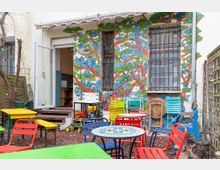 Hinterhof eines Gebäudes mit buntem Wandgemälde, das eine fantasievolle Landschaft aus Häusern, Bäumen und Straßen darstellt. Im Vordergrund befinden sich bunte Metallstühle und Tische sowie eine offene Tür, die ins Innere des Gebäudes führt.