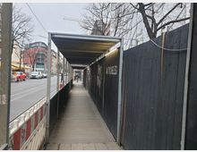 Überdachter Gehweg entlang einer Baustelle, mit einer schwarzen Holzwand auf der rechten Seite und Baugerüstabdeckungen im Hintergrund. Links neben dem Gehweg befinden sich eine Straßenbahnlinie, parkende Autos und Gebäude.