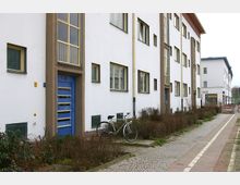 Mehrfamilienhaus mit weißer Fassade und blauen Türen im Stil der modernen Architektur, flankiert von kleinen Büschen und gepflasterten Wegen, mit einem geparkten Fahrrad im Vordergrund. Im Hintergrund sind weitere Gebäude und ein abgesperrter Bereich zu sehen.