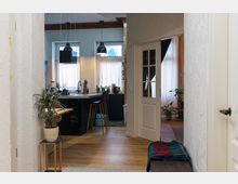 Ein Innenraum mit Blick auf eine offene Küche mit zentraler Küchentheke, schwarzen Hängelampen und Holzfußboden. Im Vordergrund steht eine Zimmerpflanze auf einem kleinen Beistelltisch, und rechts ist eine teilweise geöffnete Tür mit Glaseinsätzen zu sehen, die in einen weiteren Raum führt.