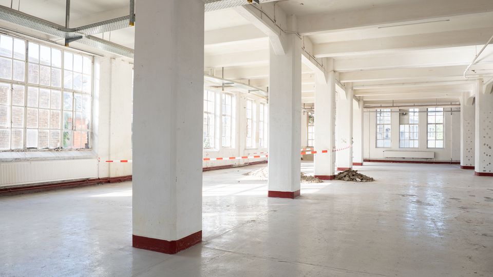 Helle, leere Industriehalle mit hohen Decken, weißen Stützpfeilern und freiliegenden Kabeltrassen. Große Sprossenfenster entlang der Außenwände lassen viel Tageslicht auf den Betonboden fallen; teilweise sind Baustellenabsperrbänder und Schutt zu sehen.