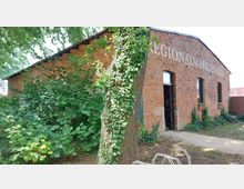 Ziegelbau eines historischen Gebäudes mit der Aufschrift „Regionalmarkthalle“ an der Fassade. Vor dem Gebäude stehen ein großer Baum mit Efeubewuchs und dichte Sträucher, daneben ein offener Eingang und Fahrradständer.