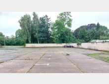 Leerer, asphaltierter Parkplatz mit vereinzelten Grasbüscheln und weißen Markierungen, umgeben von einer niedrigen, hellen Steinmauer. Im Hintergrund stehen hohe Bäume, und ein einzelnes Auto parkt rechts am Rand.