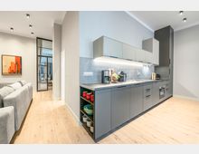 Moderne, offene Wohnküche mit langer, grauer Einbauzeile, Oberschränken und integrierten Geräten, beleuchtet durch Einbauspots und Unterbauleuchten. Heller Holzboden und angrenzender Wohnbereich mit grauem Sofa sowie Glastür zum Flur sind sichtbar.