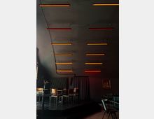 Ein dunkler Innenraum mit einer kleinen Bühne, umgeben von Holztischen und Stühlen. Von der Decke hängen rote und orangefarbene Leuchtstäbe, die den Raum beleuchten, und an der Wand befinden sich ein Heizkörper, ein Vorhang und ein Gemälde.