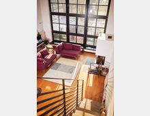 A modern living room with large floor-to-ceiling grid-patterned windows, allowing natural light to fill the space. The room features wooden flooring, a pair of magenta sofas, a patterned rug, and a contemporary fireplace near a wooden chair, viewed from the top of a staircase with metal railings.