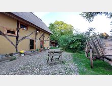 Fachwerkhaus mit Strohdach, umgeben von einem gepflasterten Hof, grüner Vegetation und einem hölzernen Leiterwagen. Vor dem Gebäude stehen Blumentöpfe und Gefäße, daneben einfache Holzbänke und ein kleiner Gartentisch.