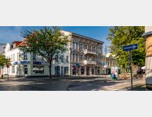 Straßenansicht einer kleinen Stadt mit mehrstöckigen Gebäuden; rechts ein historisches Gebäude mit dekorativer Fassade und einem kleinen Balkon, links ein modernes Eckgebäude mit Geschäften im Erdgeschoss. Im Vordergrund Straßenschilder und Bäume, im Hintergrund Cafétische unter Bäumen.