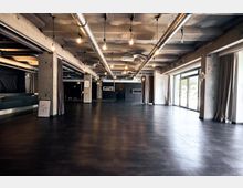 Großer, offener Raum in einem industriellen Stil mit sichtbaren Betonwänden, freiliegenden Lüftungsrohren an der Decke und dunklem Bodenbelag. Große Fensterfronten auf der rechten Seite lassen viel Licht ein, während Vorhänge an den Wänden hängen.