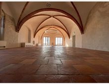 Historischer Innenraum mit einem Ziegelboden, weiß verputzten Wänden und roten Backstein-Gewölbebögen an der Decke. Am Ende des Raumes befinden sich große Fenster, die Licht hereinlassen, sowie eine Tür auf der rechten Seite.