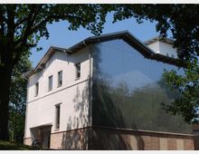 Modernes Gebäude mit weißer Fassade, großen Fenstern und einem Dach mit Sattelform. Die linke Seite besteht aus Ziegelsteinen, während an der rechten Seite eine glänzende Oberfläche reflektiert; umgeben von Bäumen und einer Wiese.