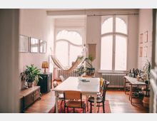 Helles Wohnzimmer mit hohen Decken und großen, geschwungenen Fenstern. Im Raum stehen ein weißer Esstisch mit Stühlen, eine Hängematte, Zimmerpflanzen sowie ein Sideboard mit Büchern und Flaschen.