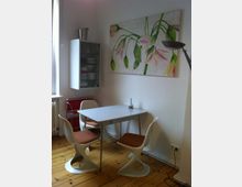 Kleine, helle Essecke mit einem rechteckigen Tisch und drei weißen Stühlen mit geschwungenem Design und braunen Sitzpolstern. An der Wand hängt ein großes Blumenbild, daneben eine schlanke, silberne Stehlampe; ein Hängeschrank mit Glastür befindet sich links, und der Holzboden ist aus hellem Parkett.