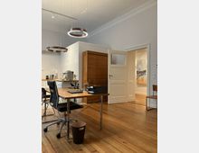 Ein helles Büro mit Holzboden, minimalistischen Möbeln und einer offenen Tür, die in einen angrenzenden Raum führt. Im Vordergrund steht ein Schreibtisch mit Stuhl, auf dem Schreibtisch befinden sich Arbeitsmaterialien; im Hintergrund eine kleine Küchenzeile und eine moderne, hängende Ringlampe.