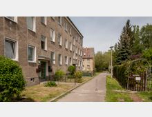 Mehrfamilienhaus mit grauer Putzfassade und mehreren weißen Fenstern, rechts daneben ein schmaler Gehweg, der von Rasenflächen, Sträuchern und Bäumen gesäumt ist. Im rechten Vordergrund befindet sich ein Torbogen mit Blumenranken, der zu einem umzäunten Garten führt.