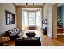 Wohnzimmer mit großen, verzierten Fensterfronten, Parkettboden und klassischer Stuckdecke. Möbliert mit schwarzen Ledersofas, einem Flachbildfernseher an der Wand und einem Beistelltisch; ein angrenzender Raum mit Schreibtisch und grünen Pflanzen ist durch eine geöffnete Tür sichtbar.