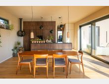 Esszimmer mit Holzfußboden und großem Esstisch aus Holz, umgeben von Stühlen mit gepolsterten Sitzflächen. Im Hintergrund eine offene Küche mit einer Holzverkleidung, Hängelampen und Zimmerpflanzen auf der Kücheninsel sowie große, bodentiefe Fenster, die viel Tageslicht hereinlassen.
