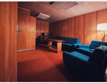 Holzvertäfelter Aufenthaltsraum mit rotem Teppichboden, abgehängter Decke mit Rasterleuchten und einem langen Holztisch in der Mitte. An den Wänden stehen blau karierte Sofas und Sessel, daneben ein kleiner TV auf einem Regal sowie eine Stehlampe.