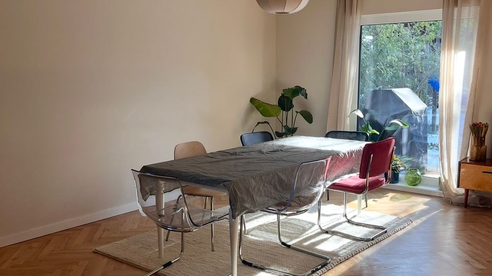 Esszimmer mit einem rechteckigen Tisch, umgeben von verschiedenen Stühlen, auf einem Teppich mit Holzboden in Fischgrätenmuster. Große Fenster mit transparenten Vorhängen lassen Tageslicht herein, neben Pflanzen im Raum. Im Vordergrund farbig gemusterte Fliesen.