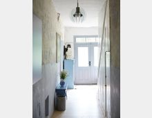 Schmaler Flur mit hellblauer Doppeltür am Ende, von natürlichem Licht beleuchtet. Links stehen ein kleiner blauer Tisch mit einer Vase voller Blumen und eine Kanne, rechts befinden sich weiß gestrichene Holzpaneele.